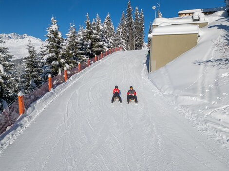Zwei Personen sitzen auf einer verschneiten Piste. Umgeben von schneebedeckten Bäumen und einem klaren blauen Himmel.
