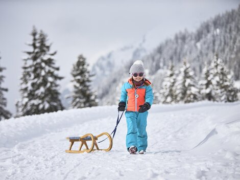 Ein Kind zieht einen Schlitten auf einem verschneiten Weg. Im Hintergrund sind verschneite Bäume und Berge zu sehen. | © TVB Paznaun - Ischgl