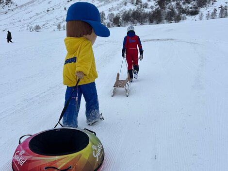 Ein Kind zieht einen Schlauchreifen auf einem verschneiten Hang. Im Hintergrund sieht man weitere Personen, die mit Schlitten unterwegs sind.