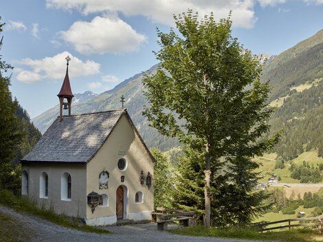 Eine kleine Kapelle umgeben von Bäumen und Bergen. Der Himmel ist partly cloudy und die Landschaft ist grün und malerisch.