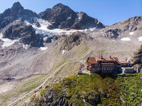 Eine Berghütte umgeben von beeindruckenden Bergen und Gletschern. Die Landschaft ist grün und bietet einen klaren Blick auf die umliegenden Gipfel.