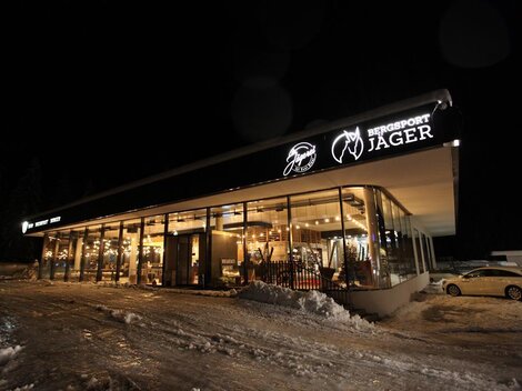 Ein modernes Sportgeschäft mit großen Fenstern, gut beleuchtet in der Nacht. Es ist von Schnee umgeben und der Geschäftsname "Bergsport Jager" ist gut sichtbar.
