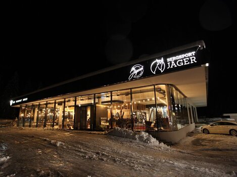 A modern sports shop with large windows, well-lit at night. It is surrounded by snow and the store name "Bergsport Jager" is clearly visible.