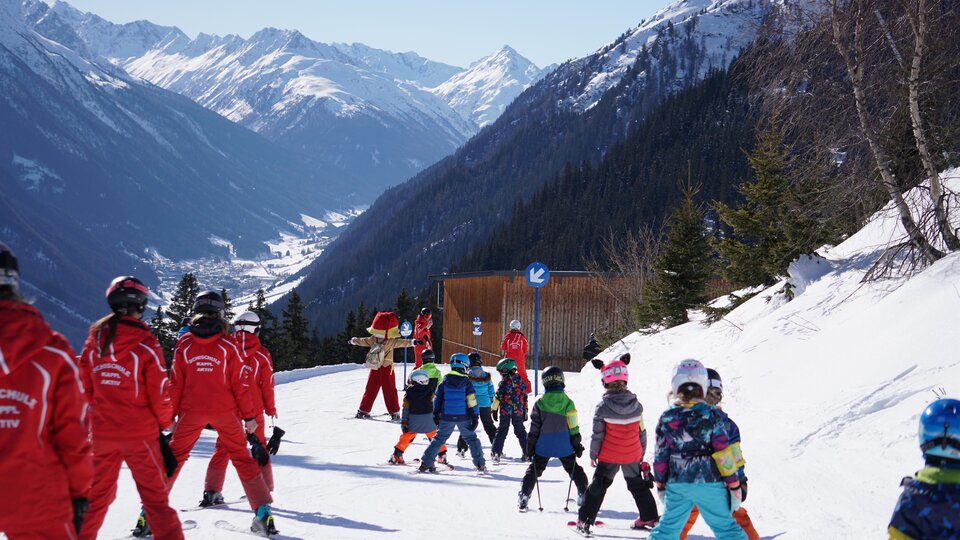 A group of children in colorful ski pants is skiing on a snowy slope. Majestic mountains can be seen in the background.