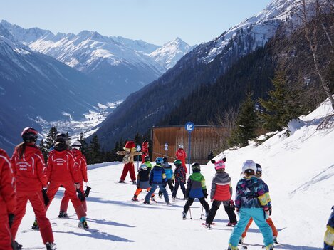 A group of children in colorful ski pants is skiing on a snowy slope. Majestic mountains can be seen in the background.