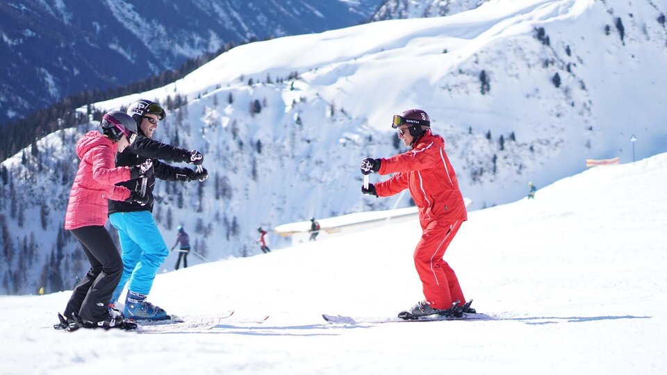 Three people in ski lessons on a snowy slope. Impressive mountains can be seen in the background.