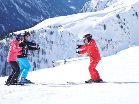 Three people in ski lessons on a snowy slope. Impressive mountains can be seen in the background.