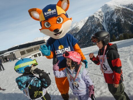 A mascot in the form of a fox stands on a snowy slope and interacts with three children. The children are wearing ski gear and are laughing.