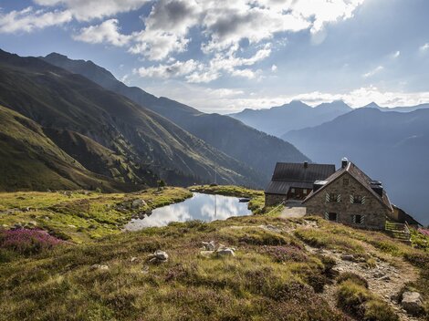 Ein malerisches Berghaus mit Blick auf grüne Wiesen und hohe Berge. Ein ruhiger Bergsee spiegelt die beeindruckende Landschaft wider.