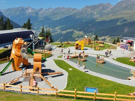 Ein Spielplatz in den Bergen mit verschiedenen Holzspielgeräten für Kinder. Im Hintergrund sind grüne Wiesen und hohe Berge zu sehen.