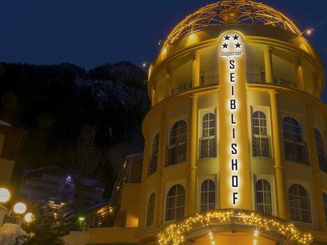 Ein hell erleuchtetes Gebäude mit dem Namen "Seiblshof" steht am Abend. Im Hintergrund sind schneebedeckte Berge sichtbar.