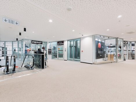 A modern interior with windows and shops. On the left side, there is an escalator and a reception desk.