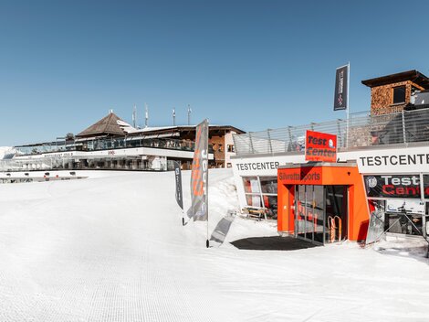 Ein schneebedecktes Skigebiet mit einem Testzentrum und einem modernen Gebäude im Hintergrund. Der Himmel ist klar und blau, ideal für Wintersportaktivitäten.
