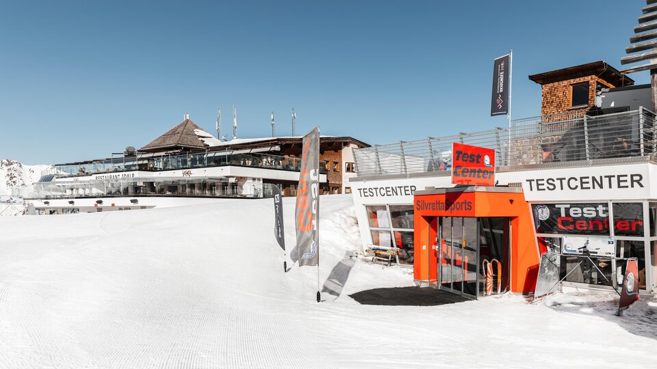 A snow-covered ski resort with a testing center and a modern building in the background. The sky is clear and blue, perfect for winter sports activities.