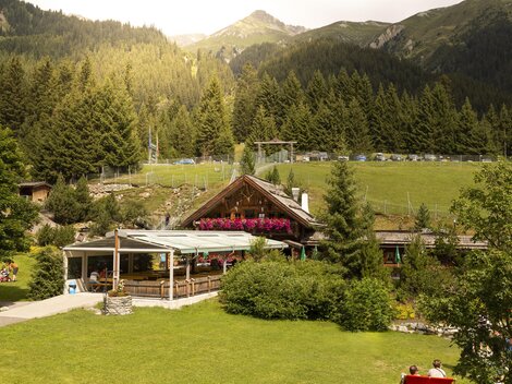 Ein gemütliches Bergrestaurant umgeben von grünen Wiesen und hohen Bäumen. Im Hintergrund sind majestätische Berge und klare Himmel zu sehen.