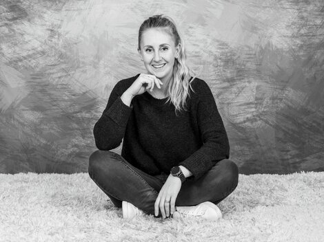 Eine junge Frau sitzt lächelnd mit gekreuzten Beinen auf einem weichen Teppich. Sie trägt einen dunklen Pullover und Jeans. | © Simone Drexel Photography