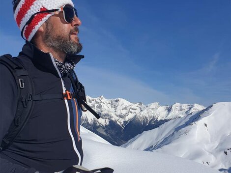 Ein Mann mit Sonnenbrille und Strickmütze steht auf einem schneebedeckten Berggipfel. Im Hintergrund sind majestätische, schneebedeckte Berge unter einem klaren blauen Himmel zu sehen.