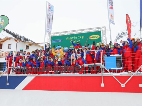 Eine Gruppe von Skifahrern in bunten Anzügen steht auf einer Tribüne. Im Hintergrund sind Schnee und Fahnen zu sehen, die für den Vitamin Cup werben.
