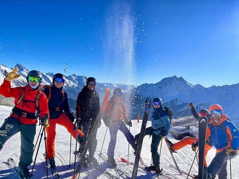 Eine Gruppe von sechs Skifahrern steht in den Bergen. Der Himmel ist klar und die Sonne scheint, während Schnee auf die Gruppe rieselt.