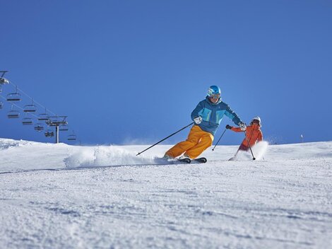 Ein Skifahrer fährt dynamisch den Hang hinunter, während ein Kind in orangefarbener Skibekleidung ihm folgt. Der Himmel ist strahlend blau und die Piste ist mit frischem Schnee bedeckt.