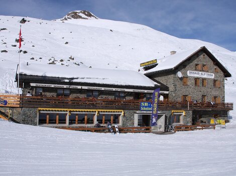 Ein gemütliches Berghaus im Schnee, umgeben von einer winterlichen Landschaft. Die Fassade ist aus Stein mit einem großen Balkon und sonnigen Terrassen.