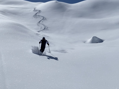 Ein Skifahrer geht durch tiefen, glitzernden Schnee unter einem klaren blauen Himmel. Im Hintergrund sind sanfte, verschneite Hügel zu sehen.