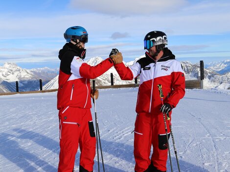 Zwei Skifahrer in roten Anzügen stehen auf einem verschneiten Gipfel. Im Hintergrund sind die majestätischen Berge sichtbar.