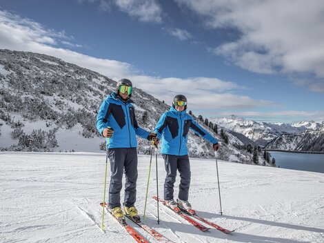 Zwei Skifahrer stehen auf einer schneebedeckten Piste. Im Hintergrund sind Berge und ein klarer Himmel zu sehen.