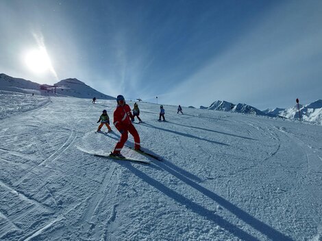 Eine Gruppe von skifahrenden Personen auf einer sonnigen Piste. Im Hintergrund sind schneebedeckte Berge zu sehen.