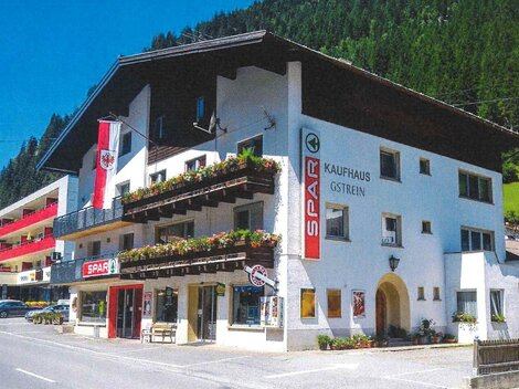 Ein kleines Geschäftshaus im alpenländischen Stil mit einem Spar-Supermarkt im Erdgeschoss. Die Fassade ist mit Blumenbalkonen und Fahnen dekoriert, umgeben von grünen Bergen.