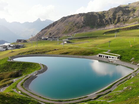 Ein malerischer Bergsee umgeben von grünen Wiesen und hohen Bergen. Die Wasseroberfläche spiegelt die Landschaft wider.