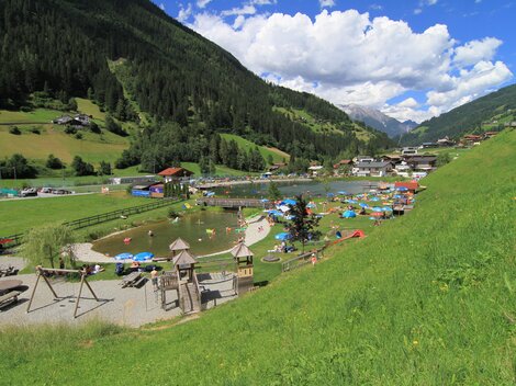 Ein malerischer Park mit einem Badesee und grünen Wiesen. Im Hintergrund sind Berge und Häuser zu sehen, während Menschen entspannen und die Sonne genießen.
