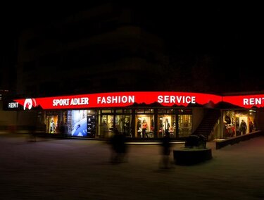 Ein Geschäft mit leuchtendem, rotem Schild, das "Sport Adler" und "Fashion Service" anzeigt. Die Szene ist nachts, und es sind verschwommene Gestalten im Vordergrund zu sehen.