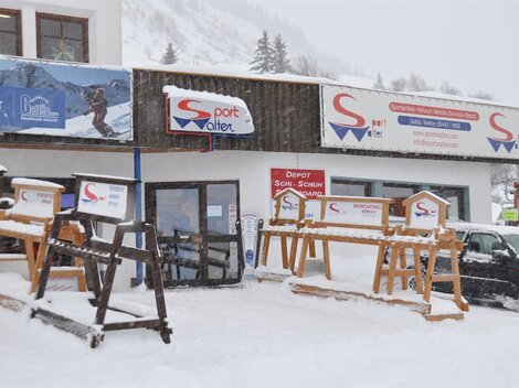 Ein Sportgeschäft im Winter mit viel Schnee vor der Tür. Die Fassade zeigt Schilder und Holzgestelle für Skiausrüstung.