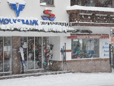 Ein Sportgeschäft mit Skiausrüstung und einer Bank im Winter. Dicke Schneeflocken fallen und die Umgebung ist verschneit.