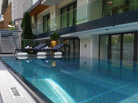 Ein elegantes Schwimmbecken mit glasklarem Wasser und modernen Liegen. Im Hintergrund ist ein stilvolles Gebäude mit großen Fenstern und Pflanzen zu sehen.