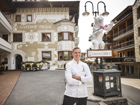A chef stands smiling in front of the Yscla Inn. In the background, the impressive building with traditional details can be seen.
