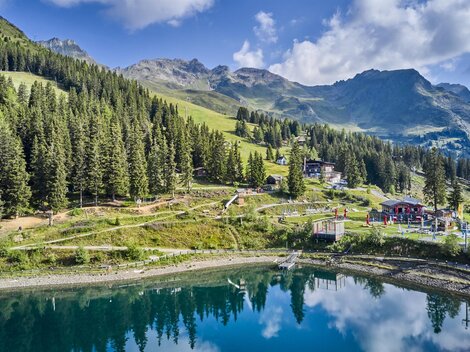Eine malerische Berglandschaft mit grünen Wäldern und sanften Hügeln. Ein ruhiger See spiegelt die Umgebung wider.