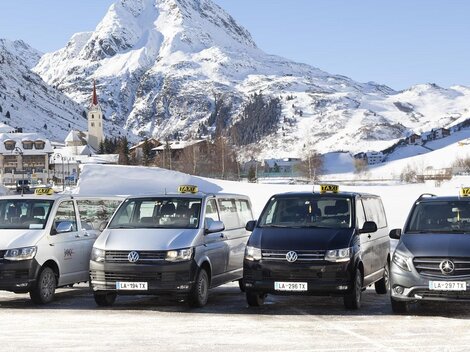 Eine Gruppe von Taxis steht vor einer schneebedeckten Berglandschaft. Im Hintergrund sind majestätische Berge und eine kleine Stadt zu sehen.