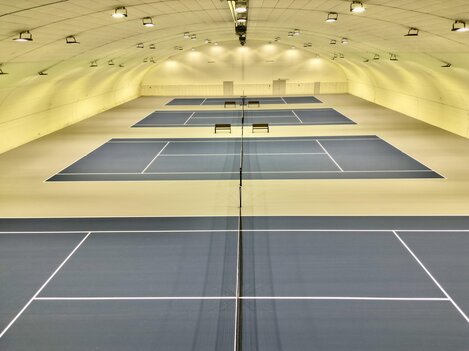 A modern tennis hall with several playing fields. The blue courts are well-lit and provide space for multiple games.