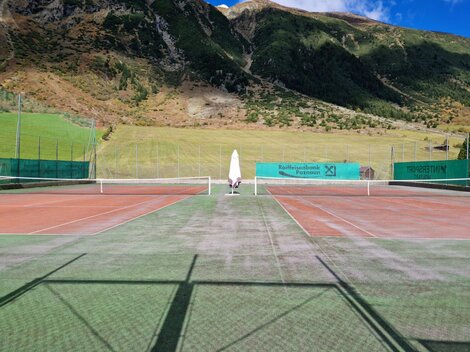 Ein Tennisplatz in einer malerischen Berglandschaft. Die Umgebung ist grün und sonnig mit klar blauen Himmel.