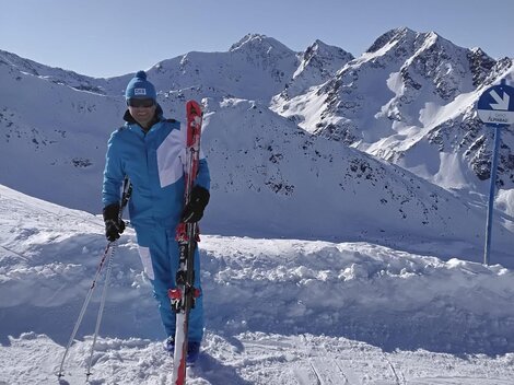 Eine Person steht im Schnee mit Skiern in der Hand. Im Hintergrund sind schneebedeckte Berge und ein klarer blauer Himmel zu sehen.
