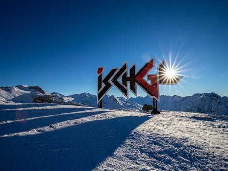 Ein wunderschöner Aussichtspunkt in Ischgl mit einem großen Schild und schneebedeckten Bergen. Die Sonne strahlt im klaren blauen Himmel.