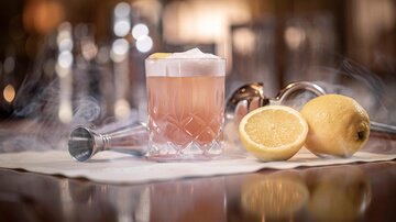Ein eleganter Cocktail in einem Glaskelch mit einer Zitronenscheibe. Daneben liegen zwei halbierte Zitronen auf einem weißen Tuch.
