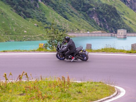 Ein Motorradfahrer fährt auf einer kurvenreichen Straße entlang eines klaren Sees. Die grüne Landschaft um ihn herum schafft eine beeindruckende Kulisse.
