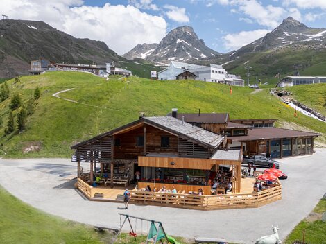 Ein modernes Holzhaus in den Bergen, umgeben von grünen Wiesen und schroffen Gipfeln. Der Himmel ist blau mit einigen weißen Wolken.