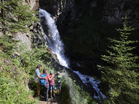 Eine Gruppe von Menschen steht an einem Wasserfall in der Natur. Umgeben von Bäumen und üppiger Vegetation genießen sie die Aussicht.