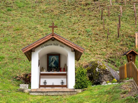 Eine kleine Kapelle mit einem Kreuz und einem Bild der Jungfrau Maria. Sie steht inmitten einer grünen Wiese.