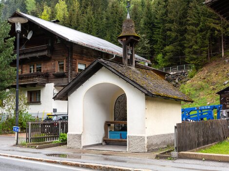 Eine kleine, weiße Kapelle mit einem Dachreiter steht an der Straße. Im Hintergrund sind typische alpine Holzhäuser und Bäume zu sehen. | © Bernhard Gruber – Photography