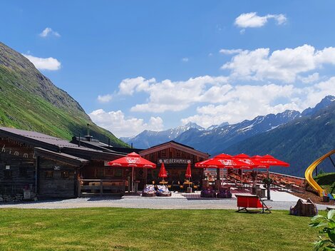 Ein gemütliches Gasthaus in den Bergen mit roten Sonnenschirmen. Im Hintergrund sind grüne Wiesen und majestätische Berge zu sehen.